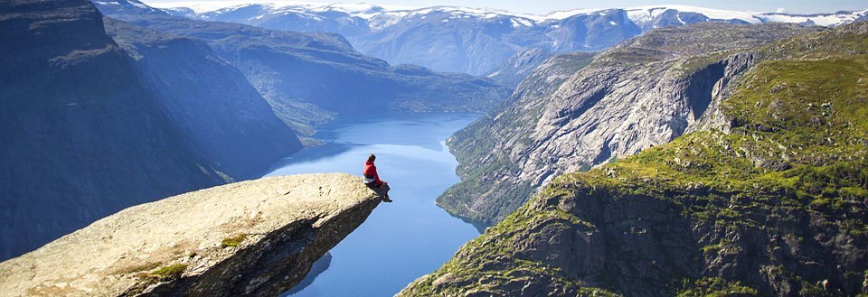 En person sidder på kanten af en fjeldklippe med udsigt over en fjord og bjerge.