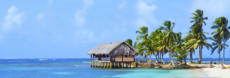 En stråtækt hytte bygget over vand omgivet af palmetræer på en tropisk strand.