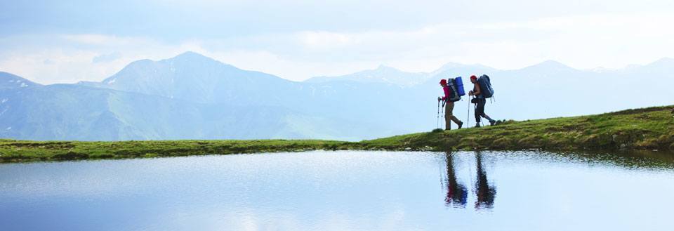 To vandrere med rygsække går ved en bjergsø med en storslået udsigt over bjergene.