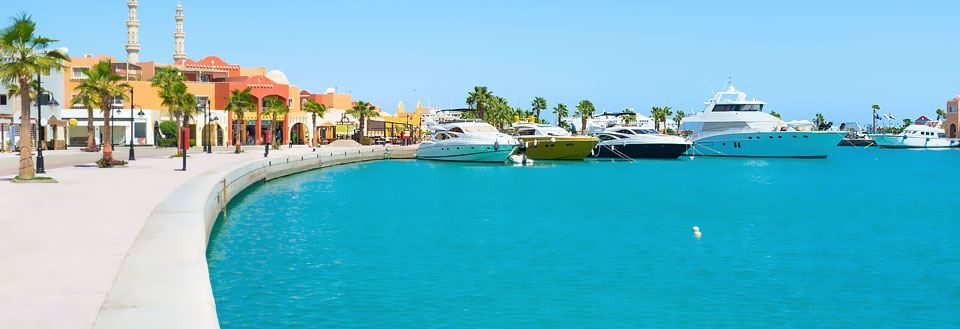 Billede af en solrig havnepromenade i Hurghada med luksusyachter fortøjet langs kajen foran farverige bygninger.