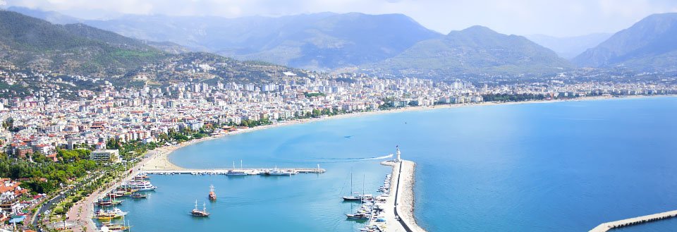 Panoramaudsigt over Alanya med klart blåt hav, en mole, både og omgivet af bjerge.