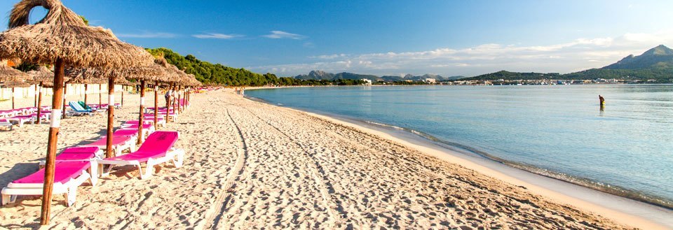 En solrig strand med parasoller, liggestole og få personer i vandet med bjerge i baggrunden.