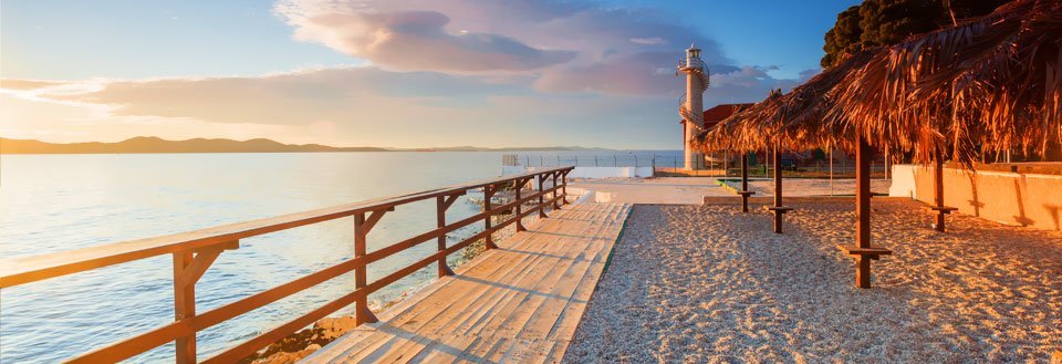 Solnedgang ved havet med en fyrtårn, strandpromenade, og en parasol-strøet strand.