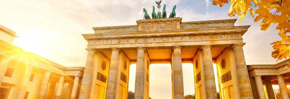 Billedet viser Brandenburger Tor i Berlin badet i solens gyldne lys med efterårsblade i forgrunden.