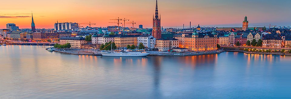 Billedet viser en panoramisk udsigt over Stockholm ved solnedgang med silhuetter af bygninger og kirkespir.