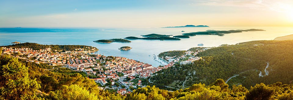 Panoramaudsigt over en malerisk kystby ved solnedgang med omgivende grønne bakker og øer i det fjerne.