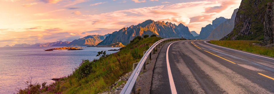 En tom vej strækker sig gennem et dramatisk landskab med fjelde ved solnedgang.