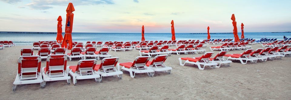 Tomme røde liggestole på en sandstrand med sammenklappede parasoller ved havet i skumringen.