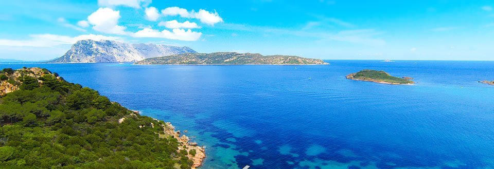 Panoramaudsigt over et krystalklart blåt hav med grønne øer og en bjergkæde i baggrunden under en blå himmel.