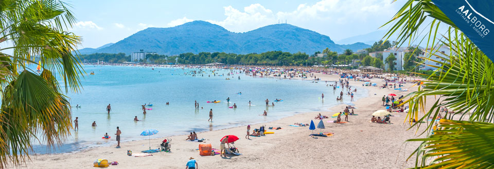 Billede af en travl strand med badegæster, farverige parasoller og bjerge i baggrunden.