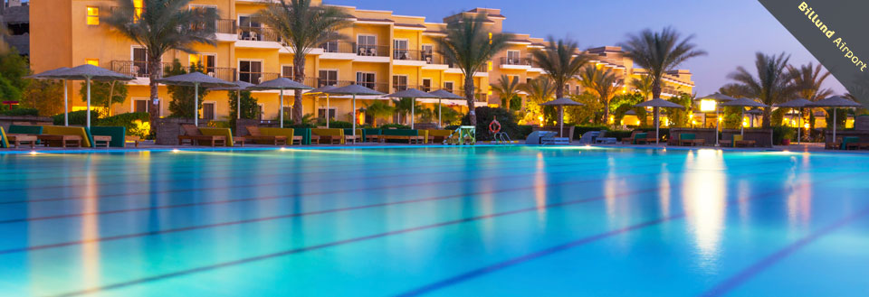 Et luksuriøst resort med swimmingpool i skumringen. Liggestole og parasoller står ved poolkanten.