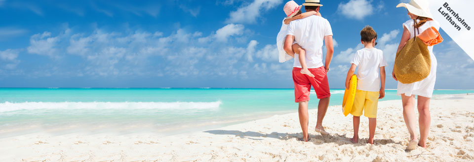 En familie med en lille dreng og pige ser på det turkisblå hav fra en solrig sandstrand.