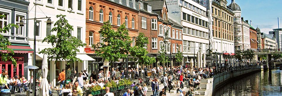 Glade mennesker nyder livet ved åen i Aarhus, bro og grønne træer under en solrig himmel.