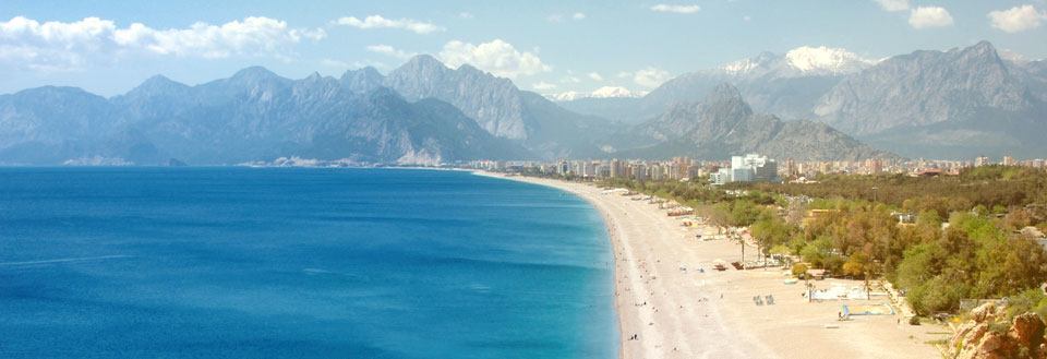 Panoramaudsigt over en blå bugt med en lang sandstrand foran og dramatiske bjerge i baggrunden.