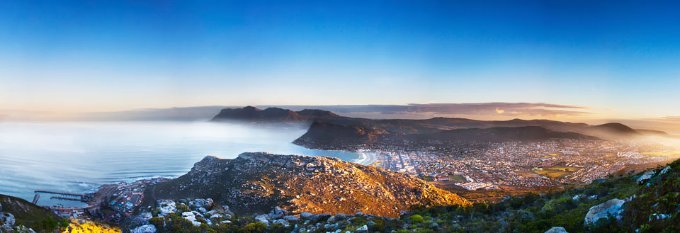 Panoramaudsigt over en kystby ved solnedgang med bjerge i baggrunden og havet i forgrunden.