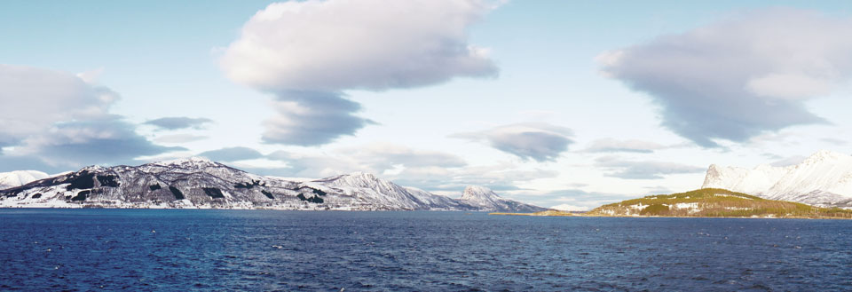 Vinterlig kystlinje med sneklædte bjerge og delvist skyet himmel.