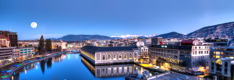 Aftenbillede af Geneve med moderne bygninger langs en flod. En klar himmel og fuldmåne ses i baggrunden.