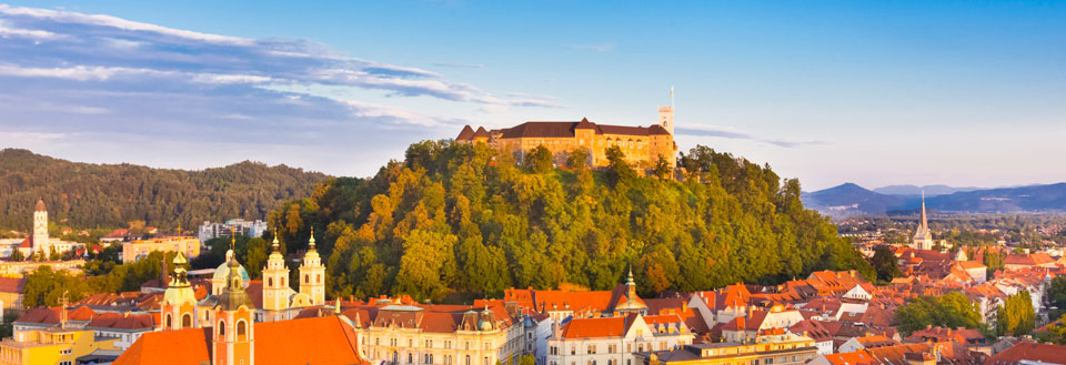 Europæisk by ved solnedgang, med historiske bygninger og et slot på en bakke.