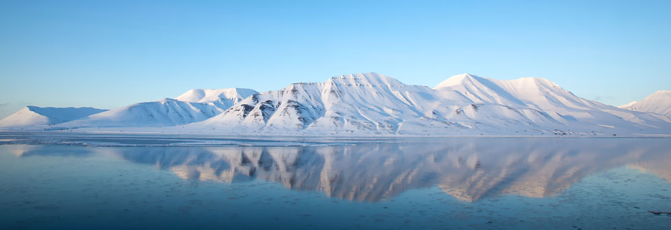 Roligt havspejl med tydelig reflektion af de sneklædte bjerge og en klar blå himmel.