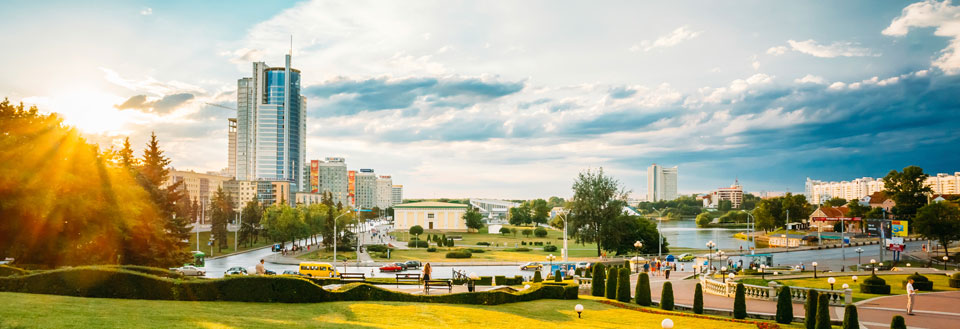 Bypark ved solnedgang med grønne plæner, høje bygninger og flod, der løber gennem.