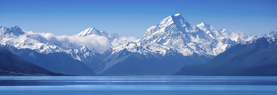 Panoramisk udsigt over en spejlblank sø i Mount Cook National Park foran majestætiske snedækkede bjerge under en klar blå himmel.