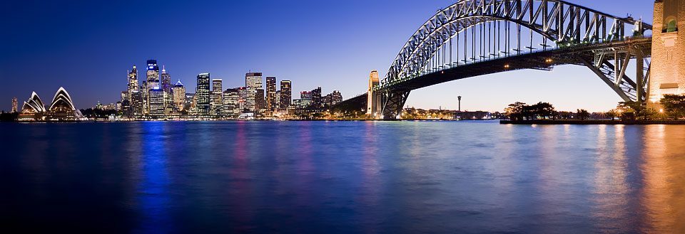 Sydneys skyline med en berømt buebro og refleksioner i vandet.