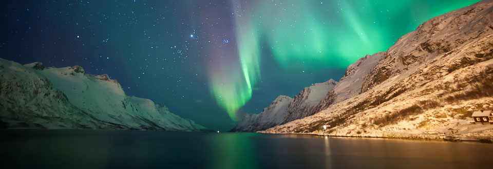 Nordlys lyser op over snebeklædte fjelde og en rolig sø med et lille hus ved bredden.