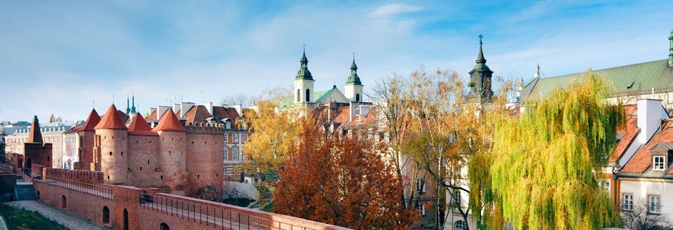 Gamle røde teglbygninger og en befæstet mur med tårne, omgivet af efterårstræer under en klar blå himmel.