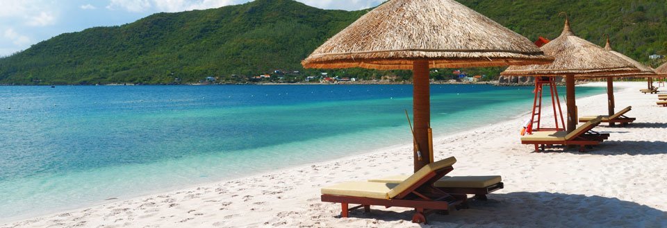 Hvid sandstrand med strandhytter og liggestole med udsigt over en klar turkis hav og grønne bjerge i baggrunden.